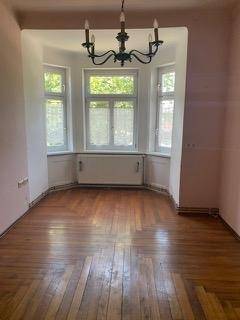 Helles Zimmer mit Erkerfenstern, Holzboden und einem klassischen Kronleuchter, bereit zur individuellen Gestaltung.