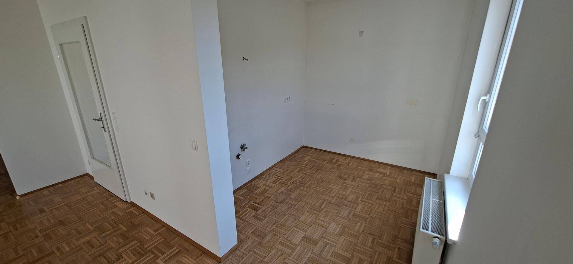 Leerer Raum mit Parkettboden und Fenster, vorbereitet für eine Küche mit sichtbaren Anschlüssen.