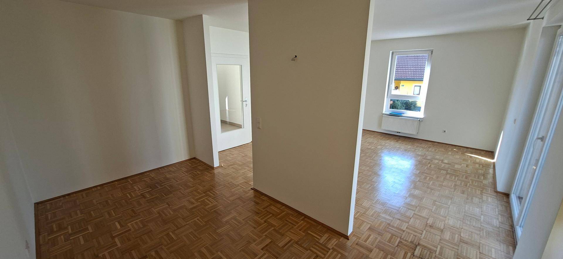 Geräumiger Wohnbereich mit Parkettboden, weißen Wänden und einem Fenster, das Tageslicht hereinlässt.