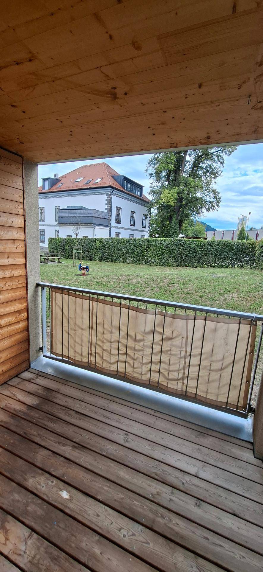 Geräumiger Balkon mit Holzboden und Blick auf einen gepflegten Garten und ein Gebäude.