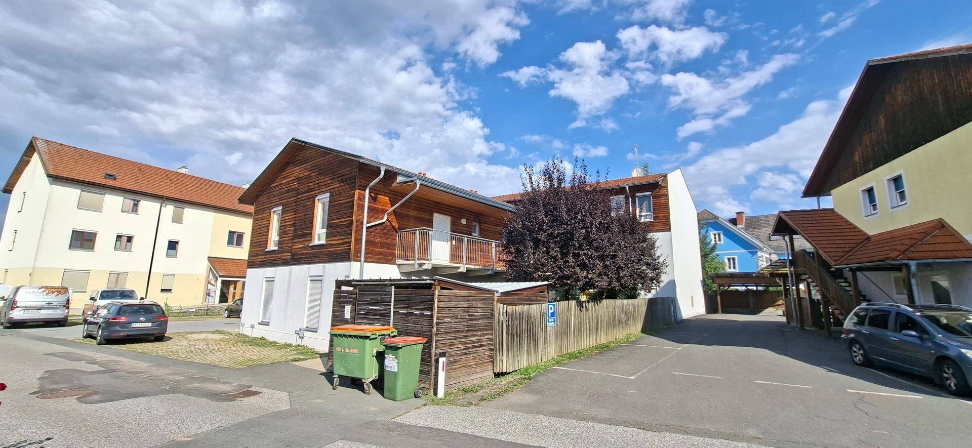 Außenansicht des Gebäudekomplexes mit Holzfassade, Parkplätzen und Mülltonnen im Vordergrund.