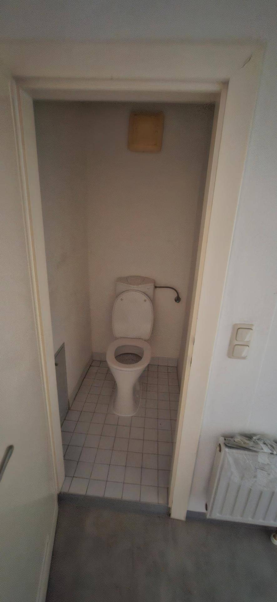 Kleines WC mit weiß gefliestem Boden, Toilette und Heizkörper in einem schlichten Raum.