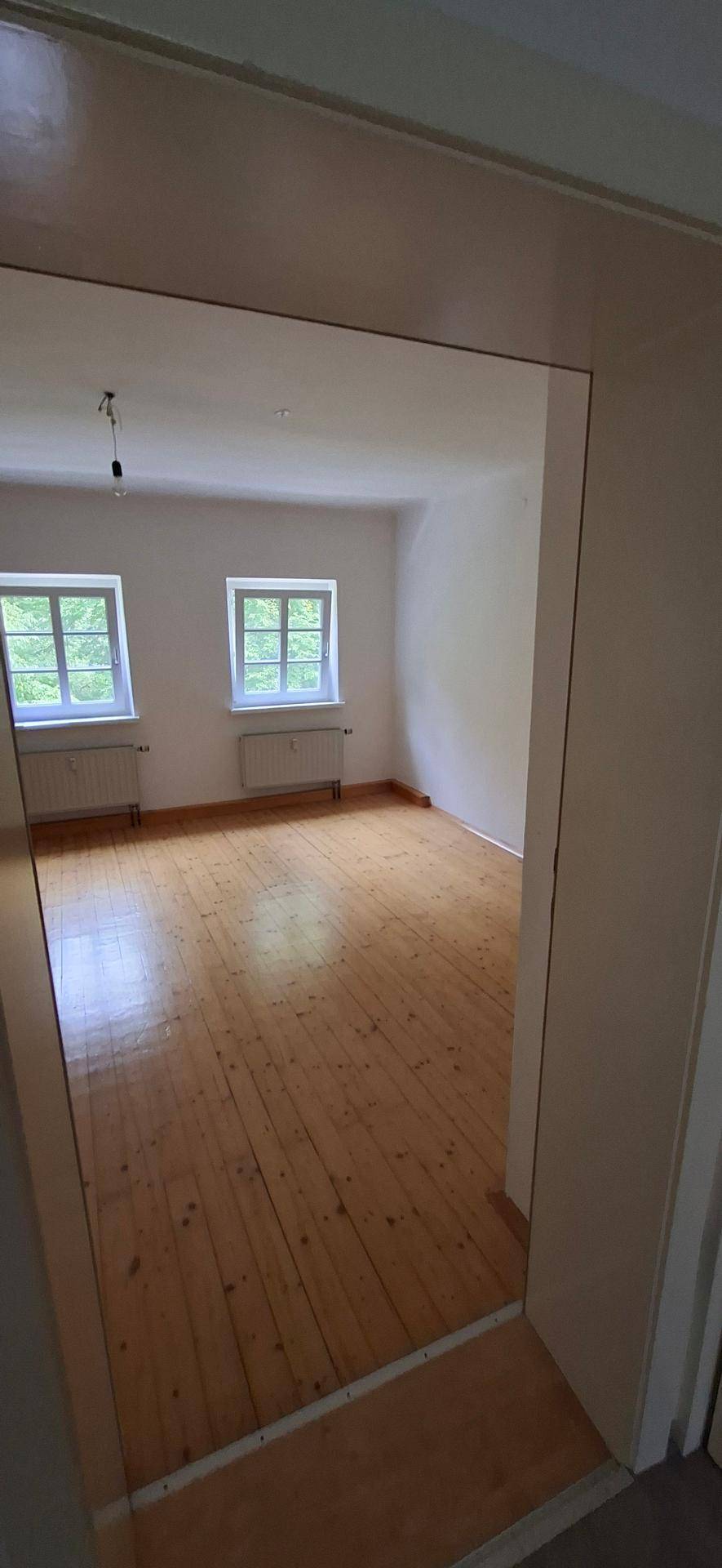 Geräumiger, heller Raum mit breiten Holzplanken und zwei Fenstern, ideal als Wohnbereich.