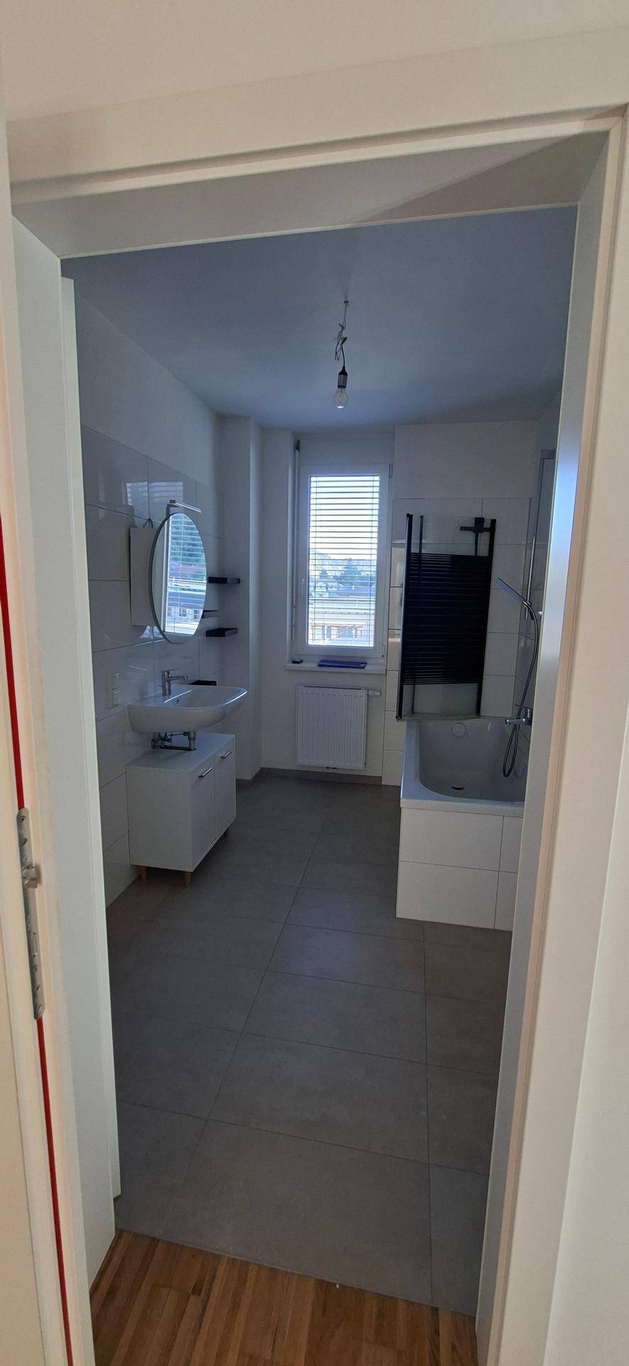 Modernes Badezimmer mit grauen Fliesen, Badewanne, Waschbecken und Fenster für natürliche Belüftung.