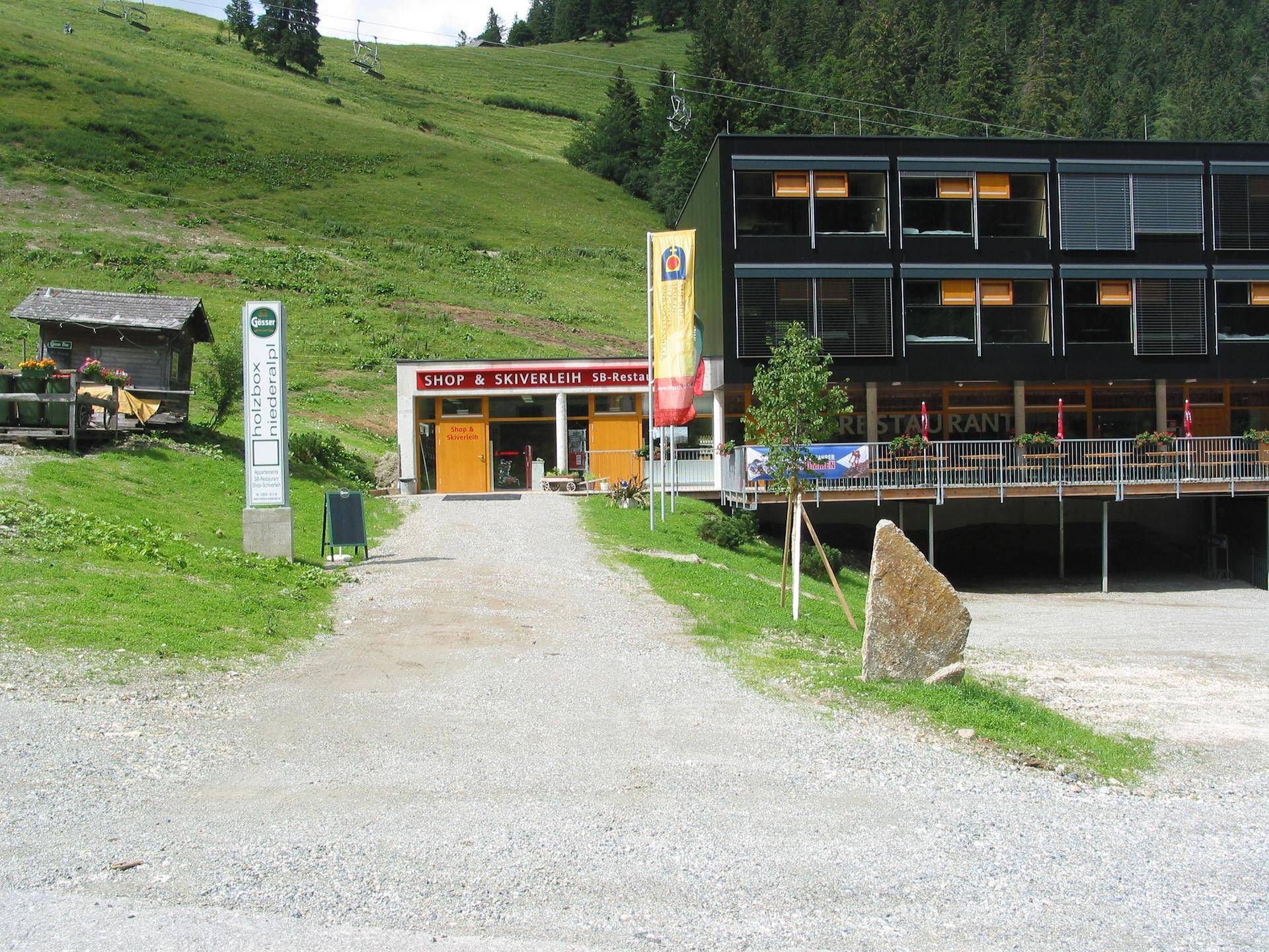 Eingangsbereich des Gebäudes mit "Shop & Skiverleih" Schild und Schotterweg in grüner Umgebung.