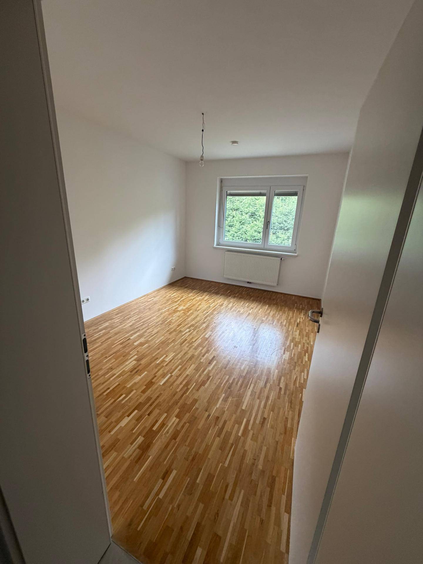 Lichtdurchflutetes Zimmer mit hellem Parkettboden und Blick auf die grüne Umgebung.