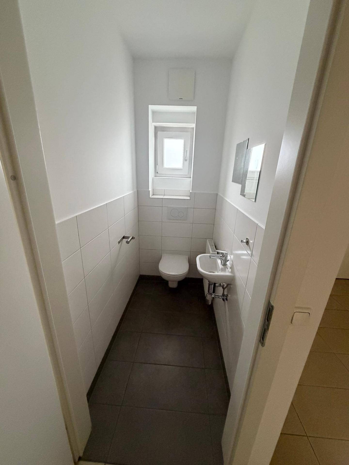 Funktionelles Gäste-WC mit weißen Fliesen, Toilette, kleinem Waschbecken und Fenster.