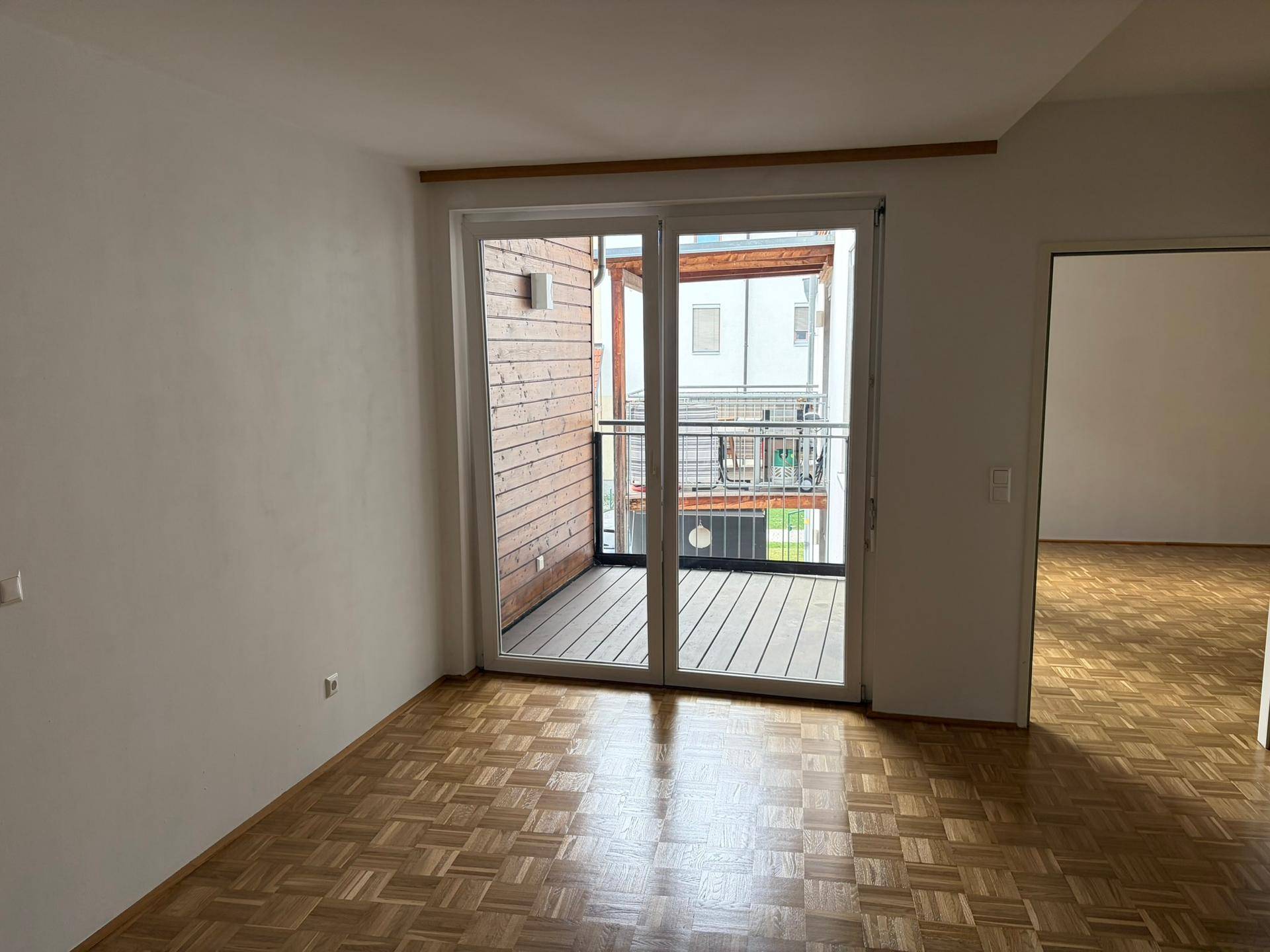 Geräumiges Zimmer mit Parkettboden und Schiebetür, die zu einem Balkon mit Blick auf die Umgebung führt.