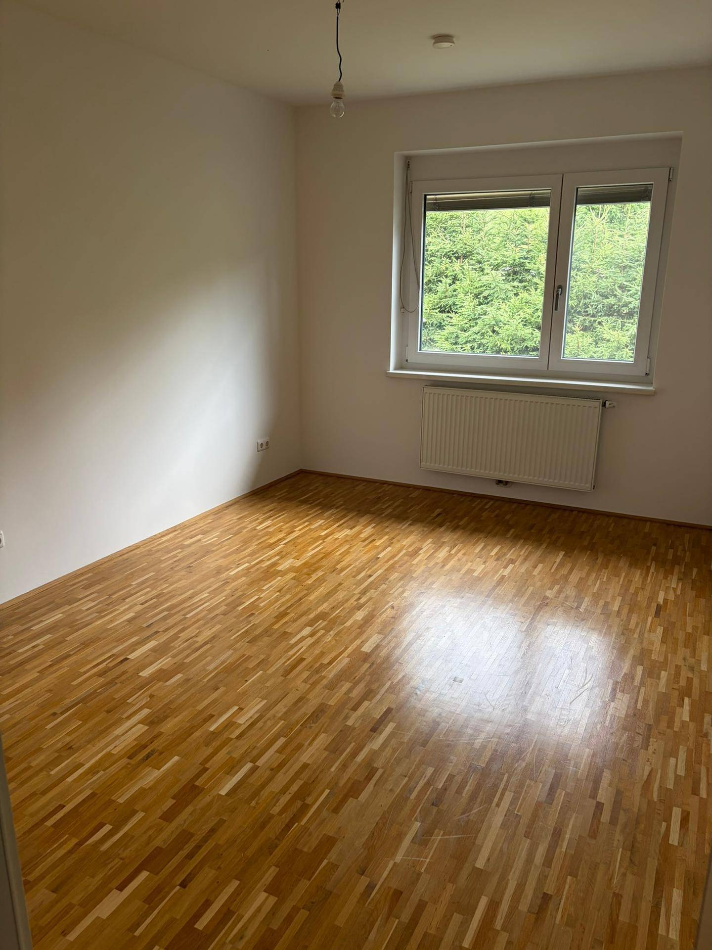 Freundliches Zimmer mit hellem Parkettboden und Fenster, das viel Tageslicht hereinlässt.