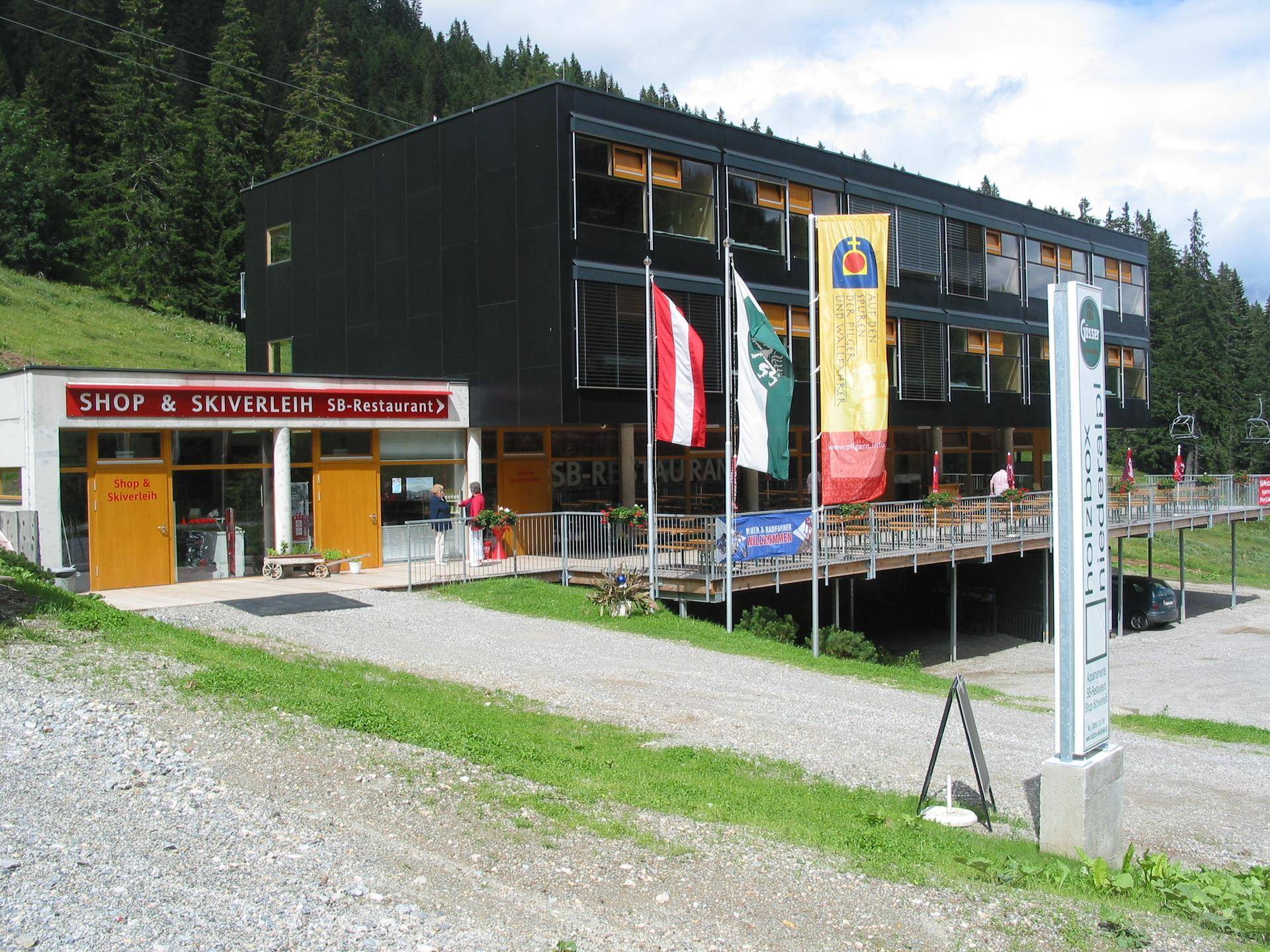 Eingangsbereich des Gebäudes mit Shop und Skiverleih sowie einer Außenterrasse in bergiger Landschaft.