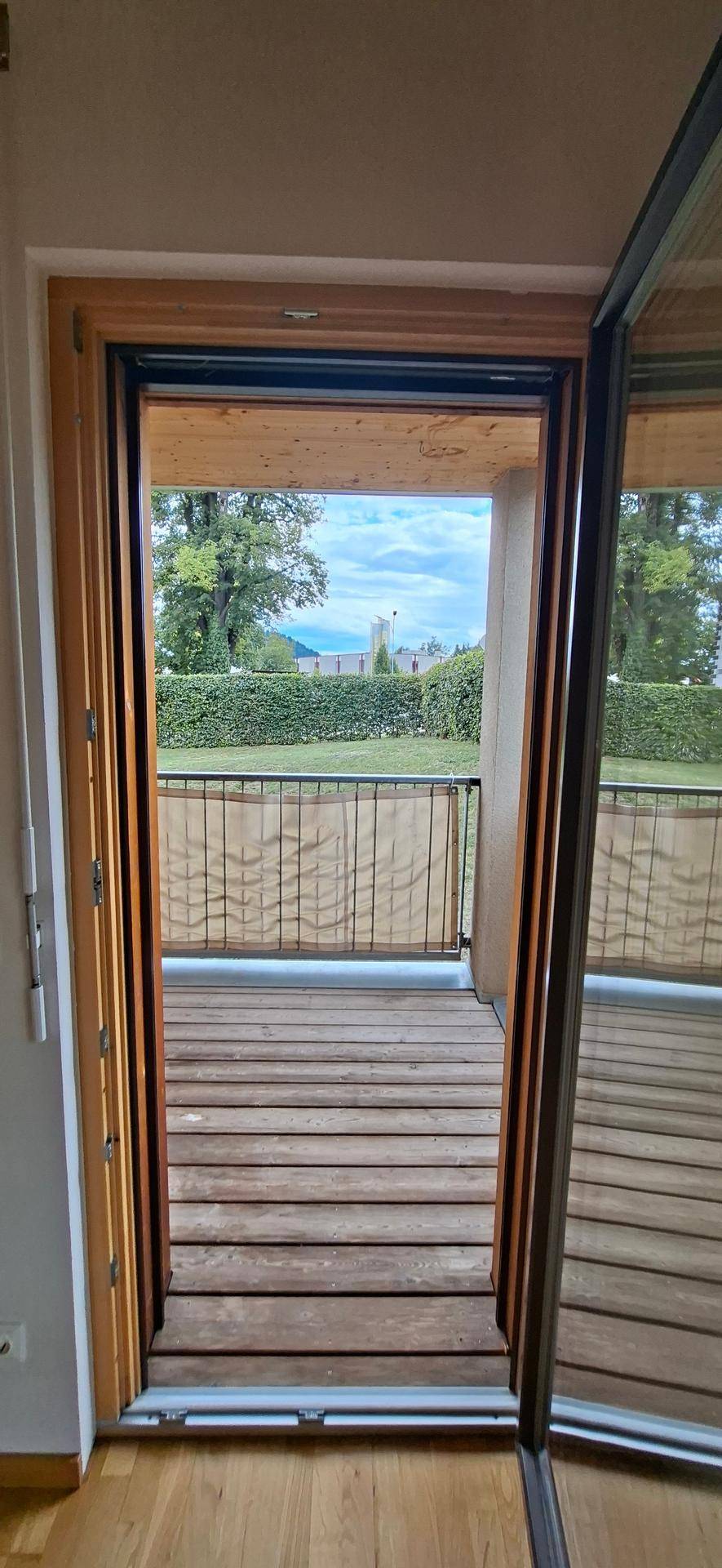 Blick vom Innenraum auf einen Holzbalcon mit Metallgeländer und grüner Umgebung.