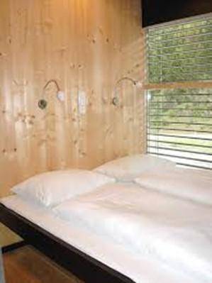 Schlafzimmer mit Holzwänden, einem Bett mit weißer Bettwäsche und einem Fenster mit Jalousien.