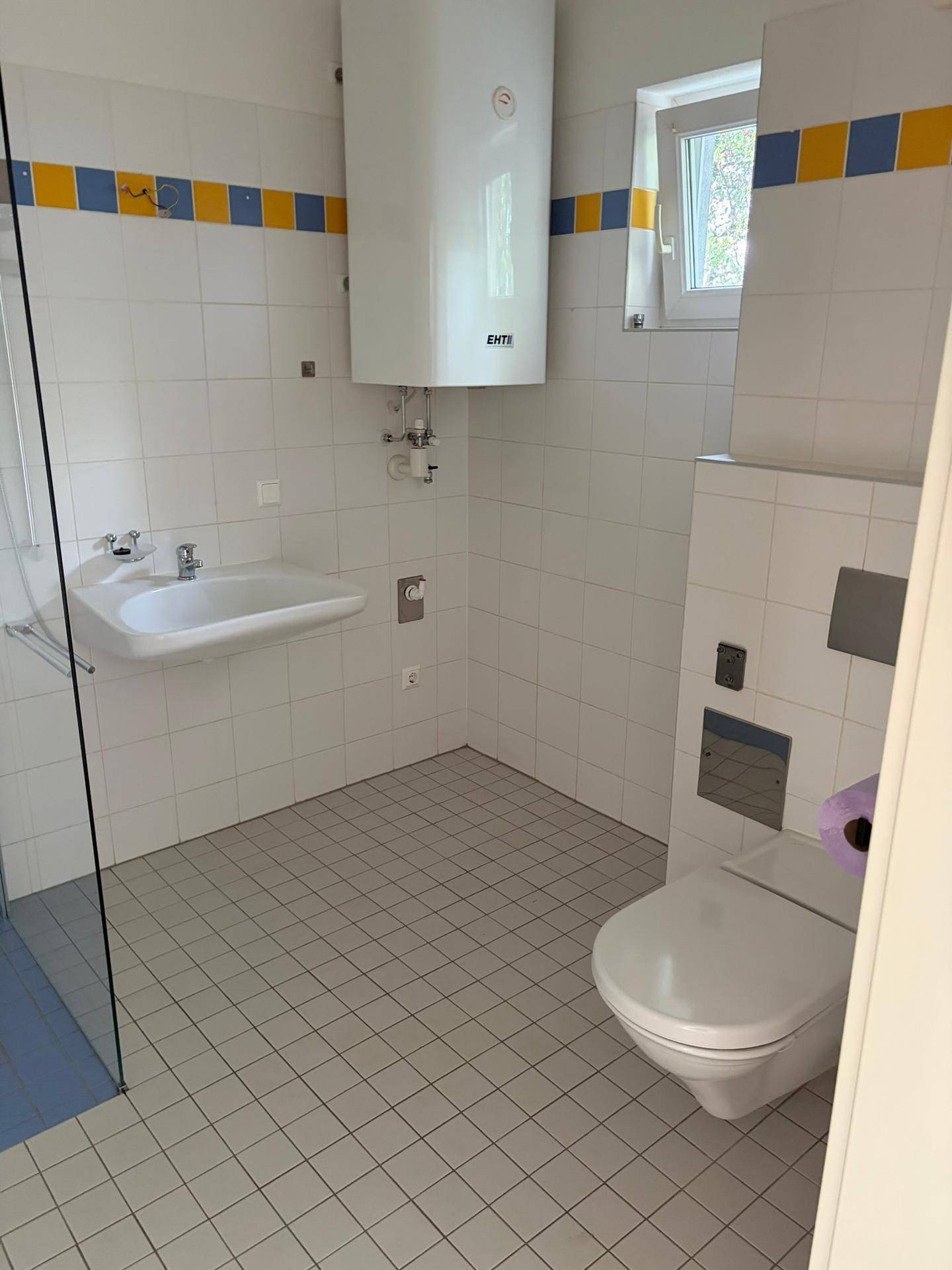 Helles Badezimmer mit WC, Waschbecken, Fenster und einem Warmwasserboiler an der Wand.
