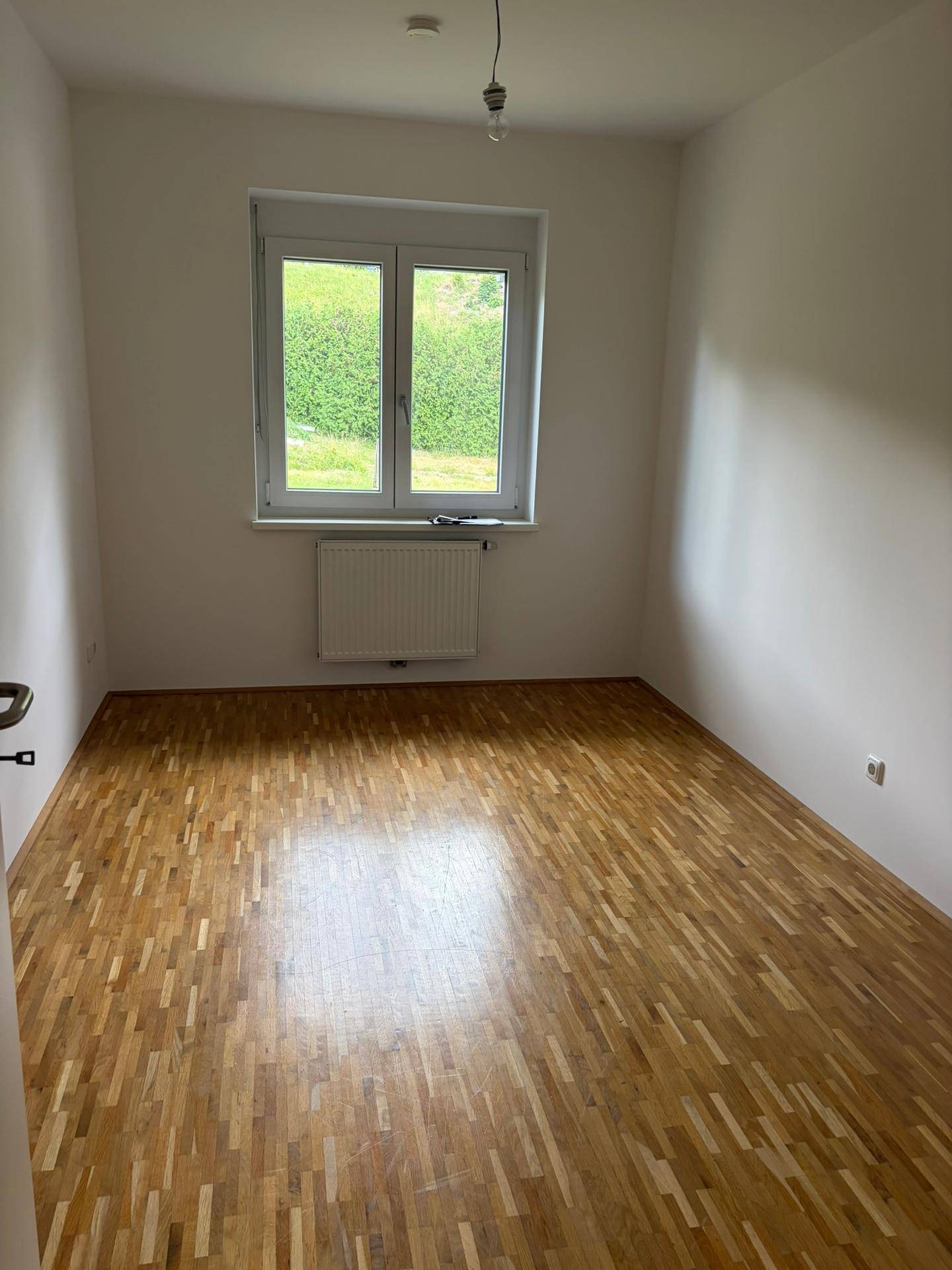 Helles Zimmer mit Parkettboden, weißen Wänden und Fenster mit Blick ins Grüne.