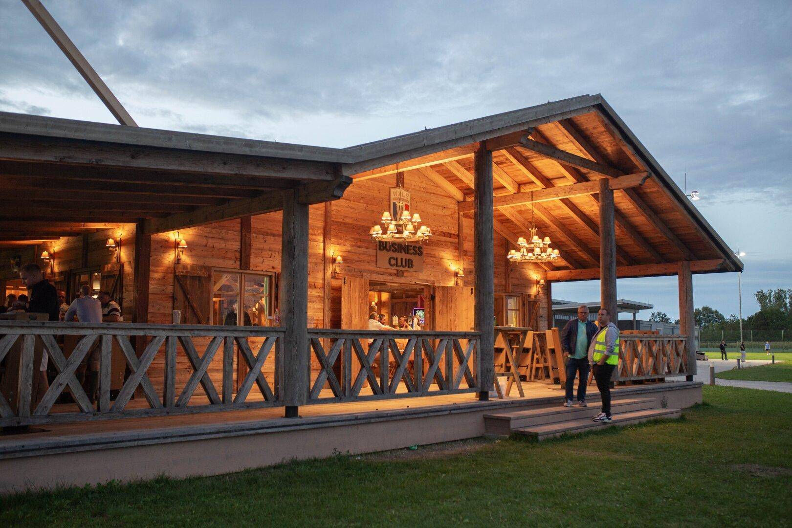 Holzgebäude mit Veranda und Kronleuchtern bei Dämmerung, umgeben von Grünflächen.