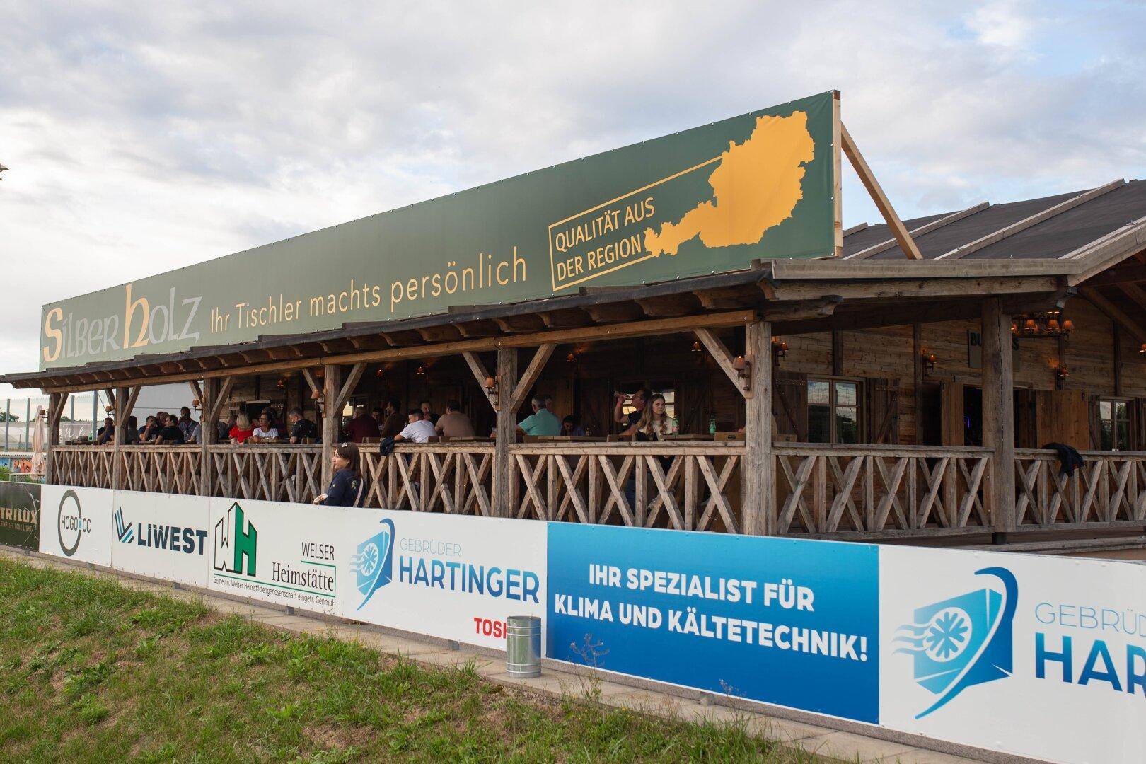Holzgebäude mit großer Veranda und Werbebannern, tagsüber unter bewölktem Himmel.