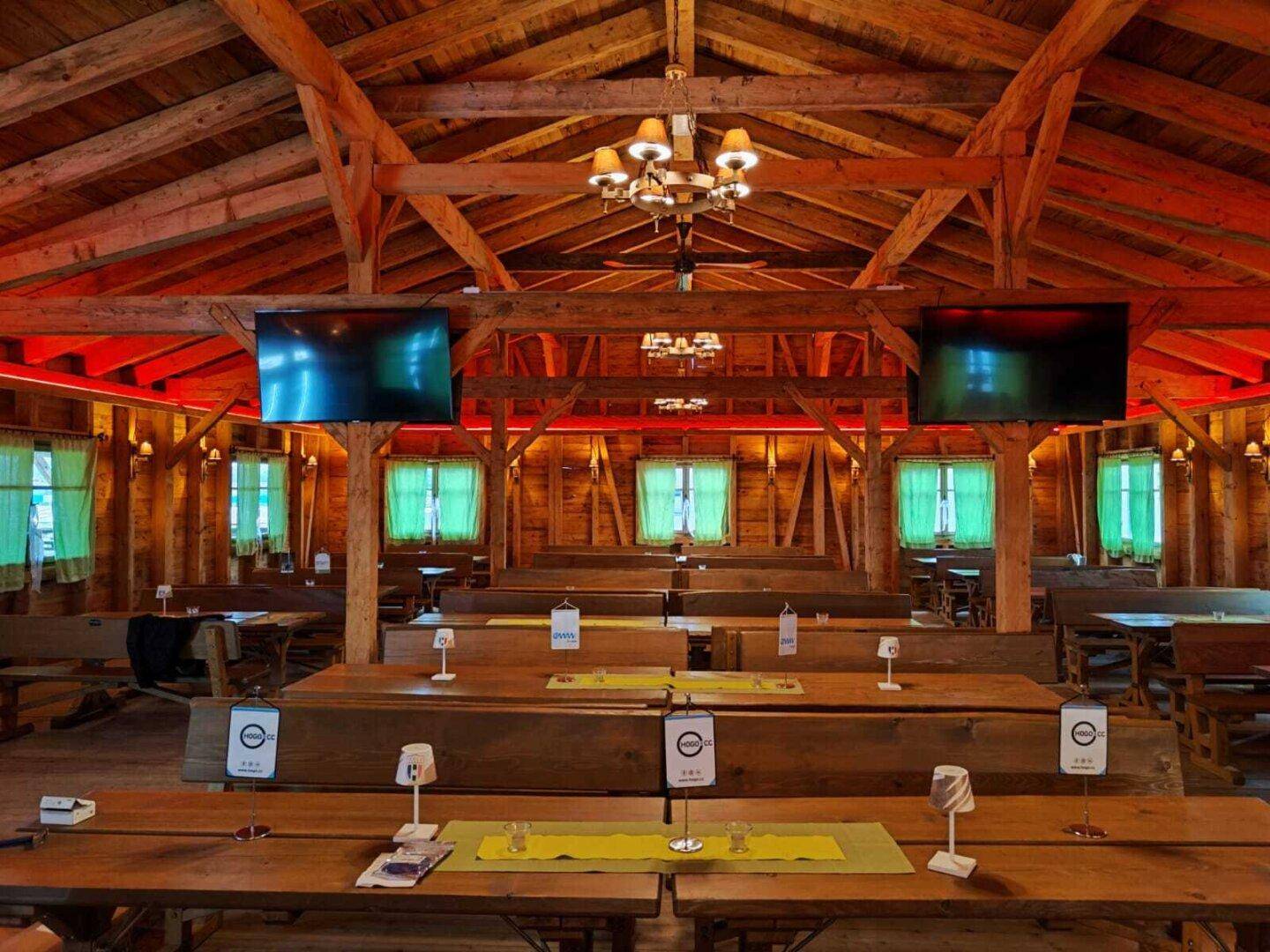 Festsaal mit langen Holztischen und Bänken, beleuchtet mit roten Lichtern und Bildschirmen.