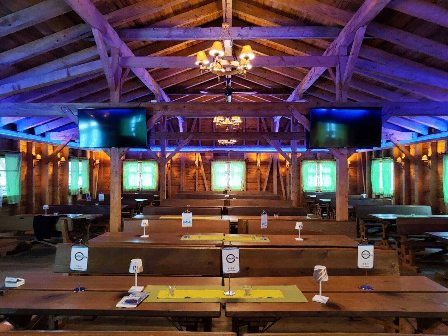 Festsaal mit langen Holztischen und Bänken, beleuchtet mit farbigen Lichtern und Bildschirmen.