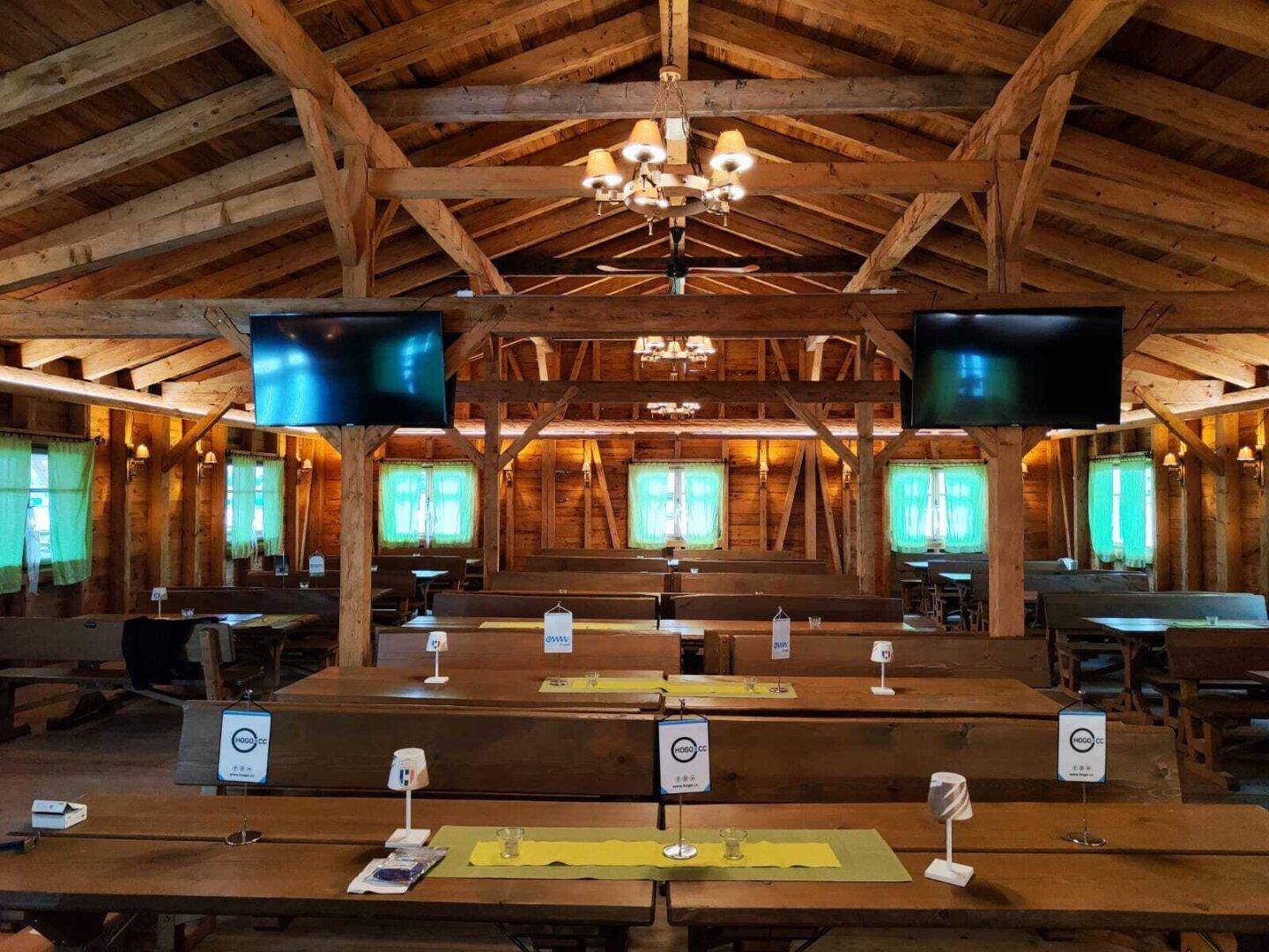 Festsaal mit langen Holztischen und Bänken, beleuchtet mit farbigen Lichtern und Bildschirmen.