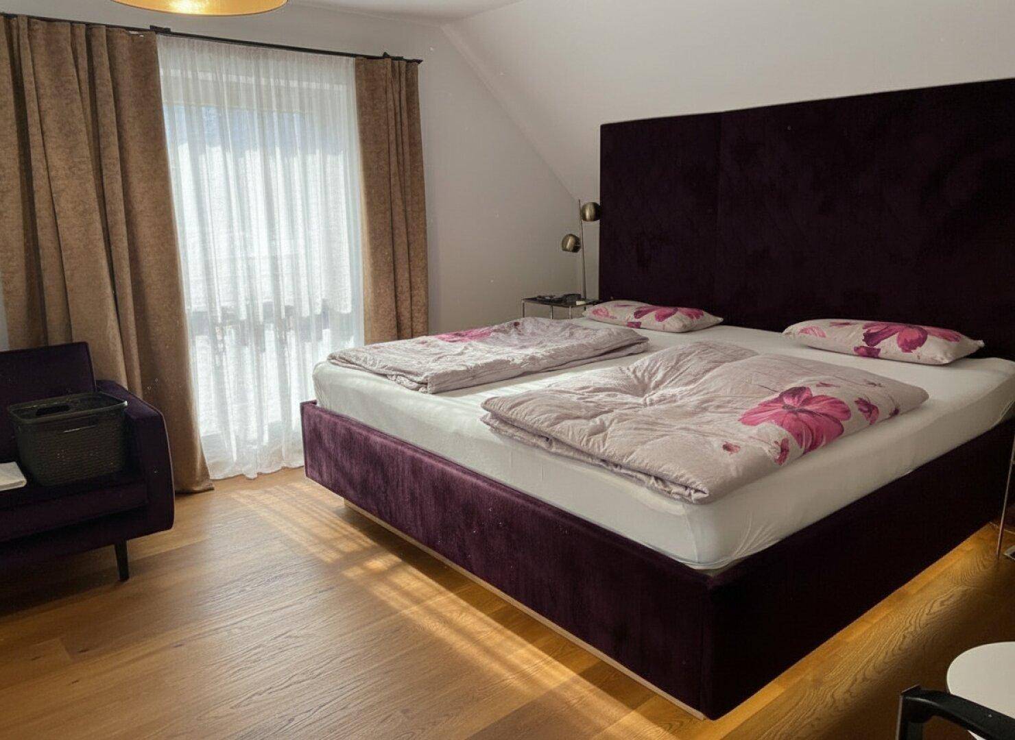 Gemütliches Schlafzimmer mit großem lila Bett, Holzfußboden und viel natürlichem Licht.