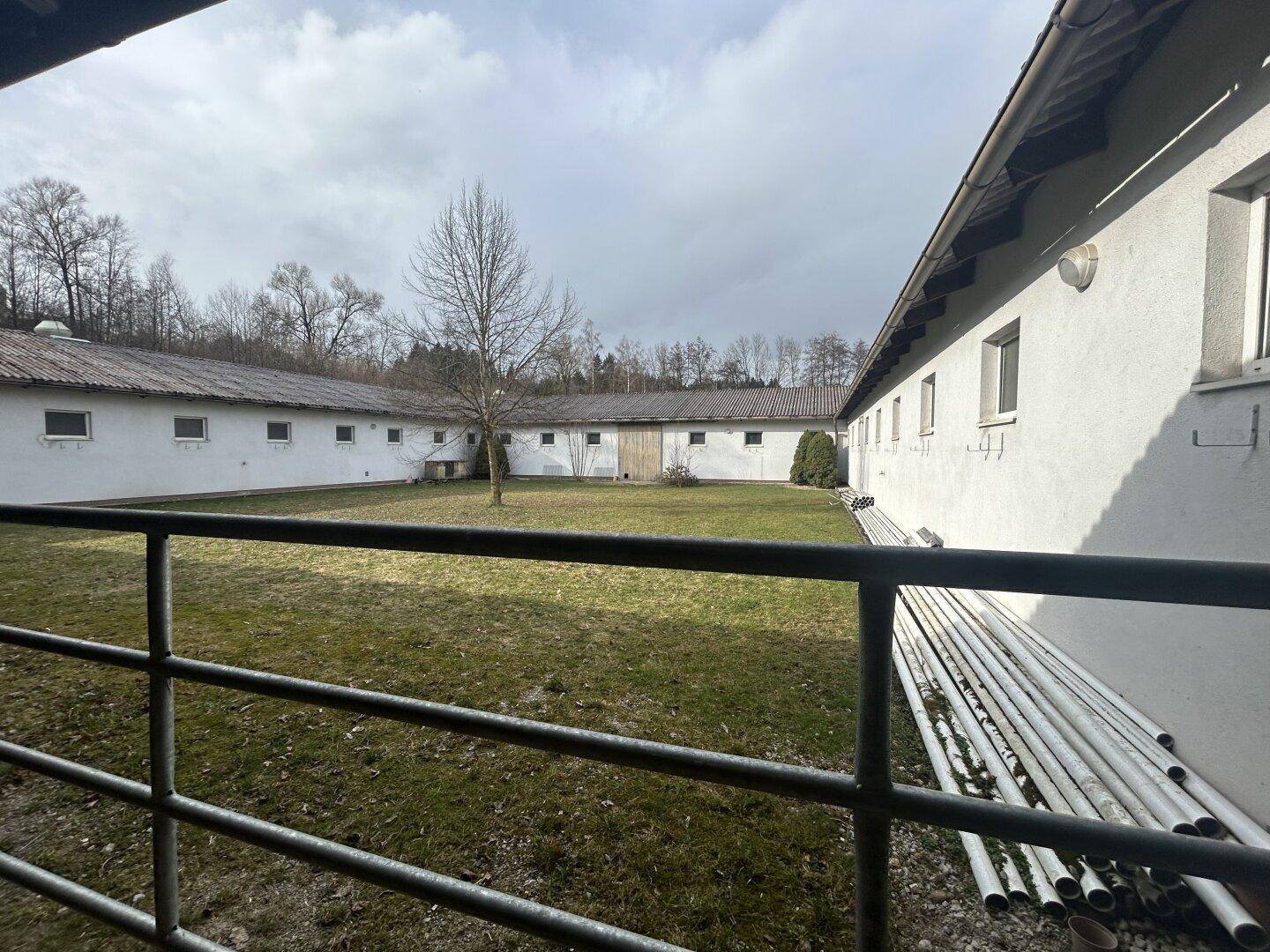 Innenhof zwischen mehreren Stallgebäuden mit einer Rasenfläche und gelagerten Rohren.