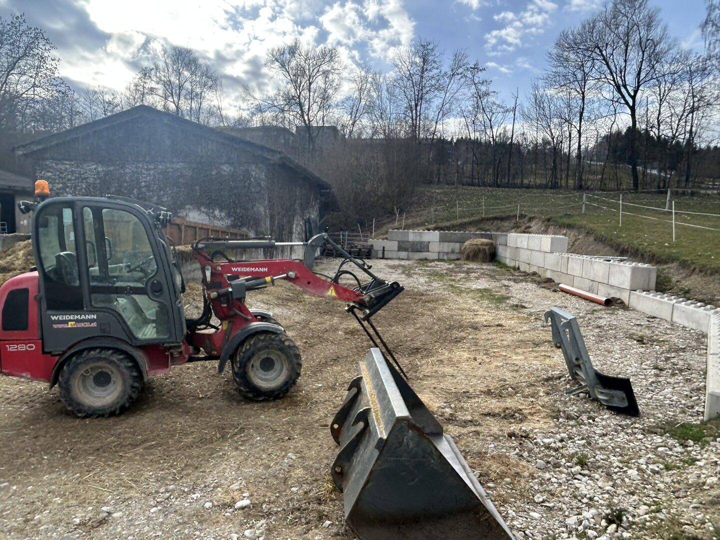 Kleiner Radlader auf einem Feldweg vor einem landwirtschaftlichen Gebäude mit Bäumen im Hintergrund.