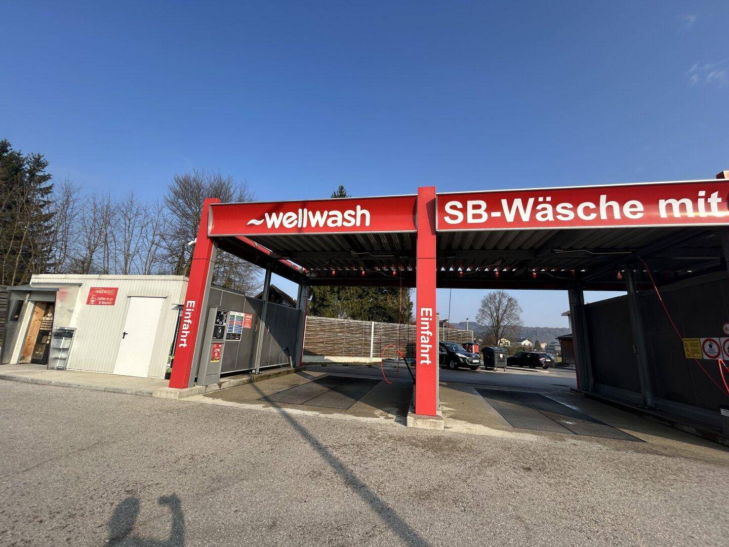 Eingangsbereich und mehrere Waschboxen einer Selbstbedienungs-Waschanlage unter blauem Himmel.
