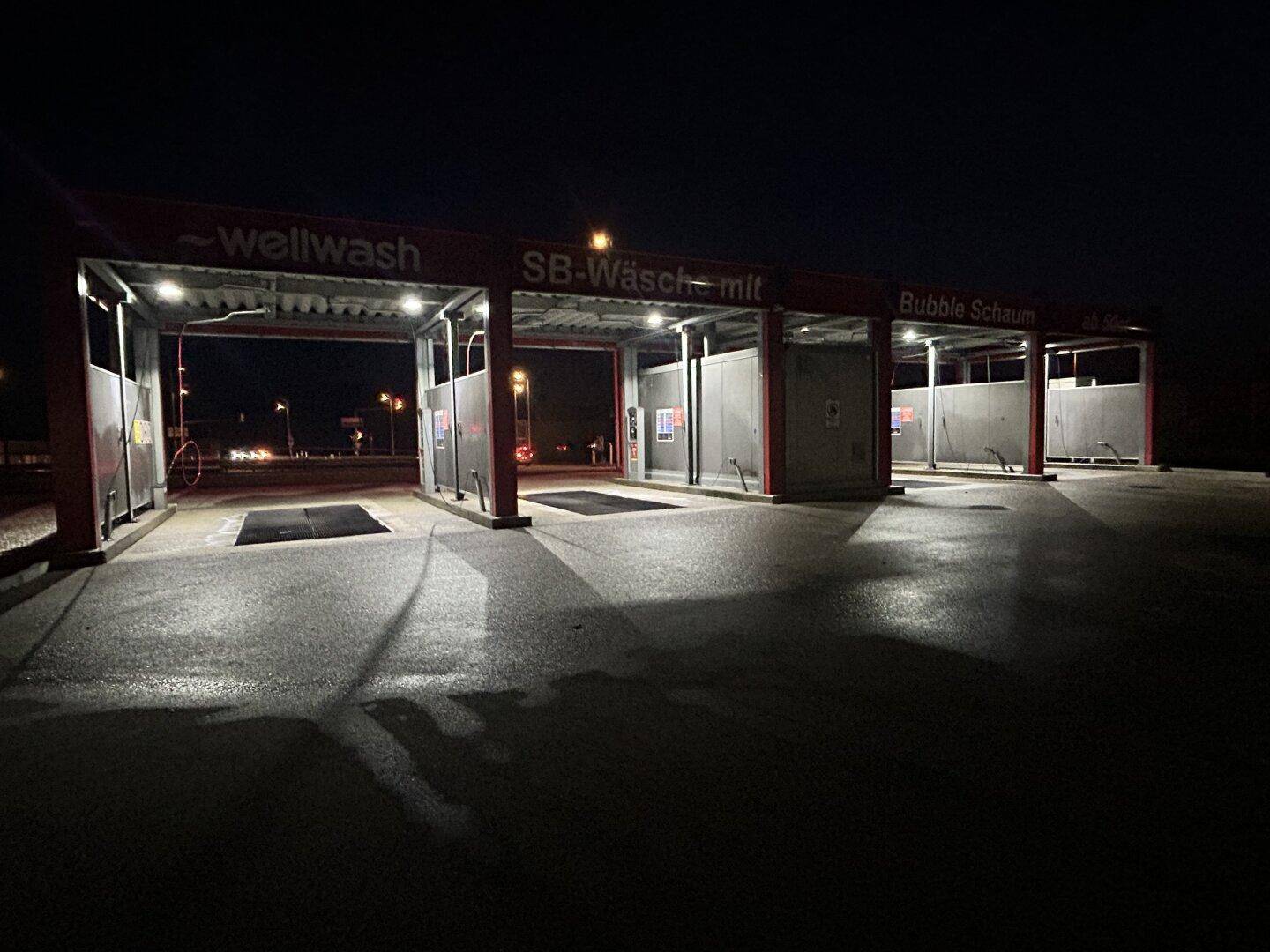 Gesamtansicht der beleuchteten SB-Waschanlage bei Nacht, mit mehreren leeren Waschboxen und dunklem Himmel.