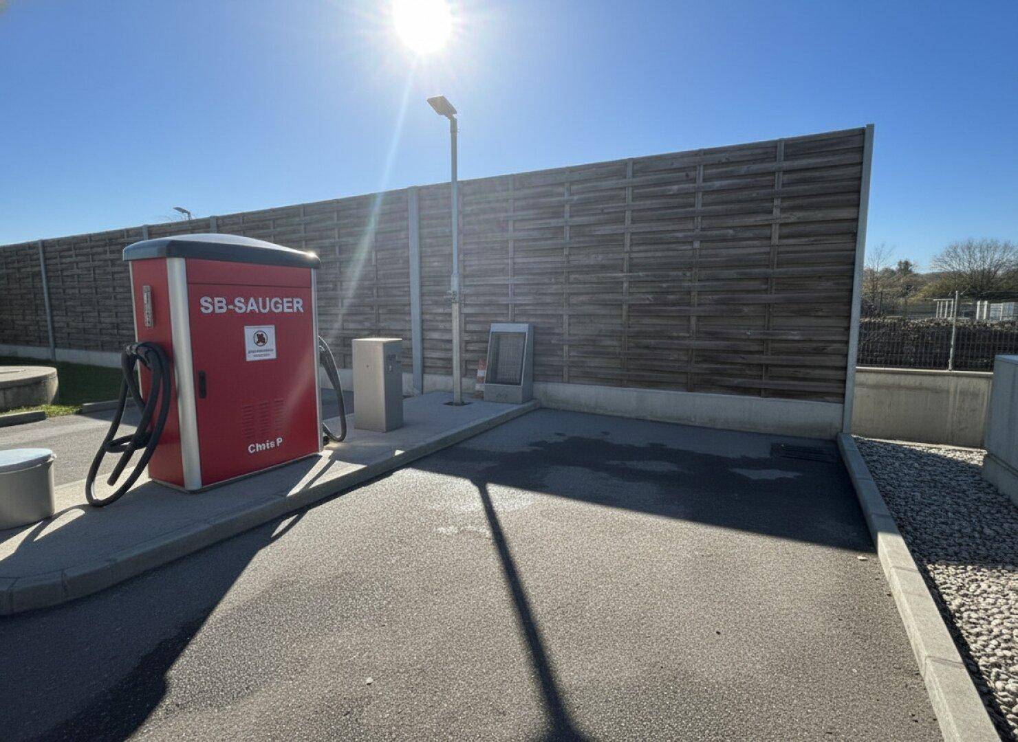 SB-Saugerstation im Freien an einem sonnigen Tag, mit einer hohen Holzwand im Hintergrund und klarem Himmel.