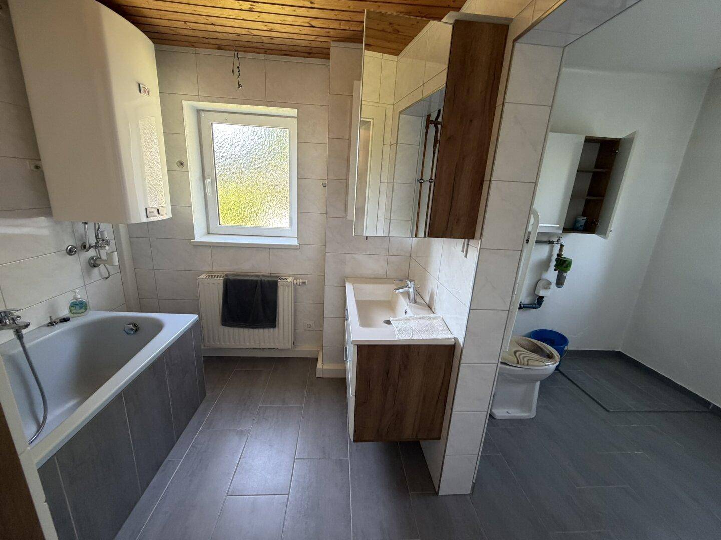 Badezimmer mit Badewanne, Waschbecken, WC und Fliesenboden unter einer Holzdecke.
