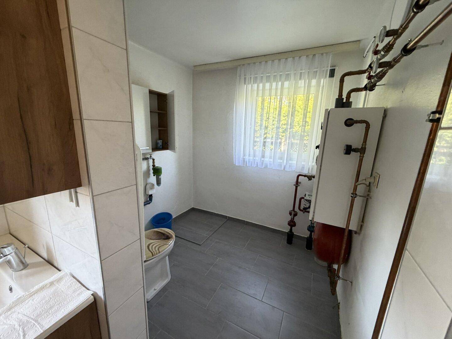 Teil des Badezimmers mit WC, Fenster und sichtbaren Installationen für Warmwasser.