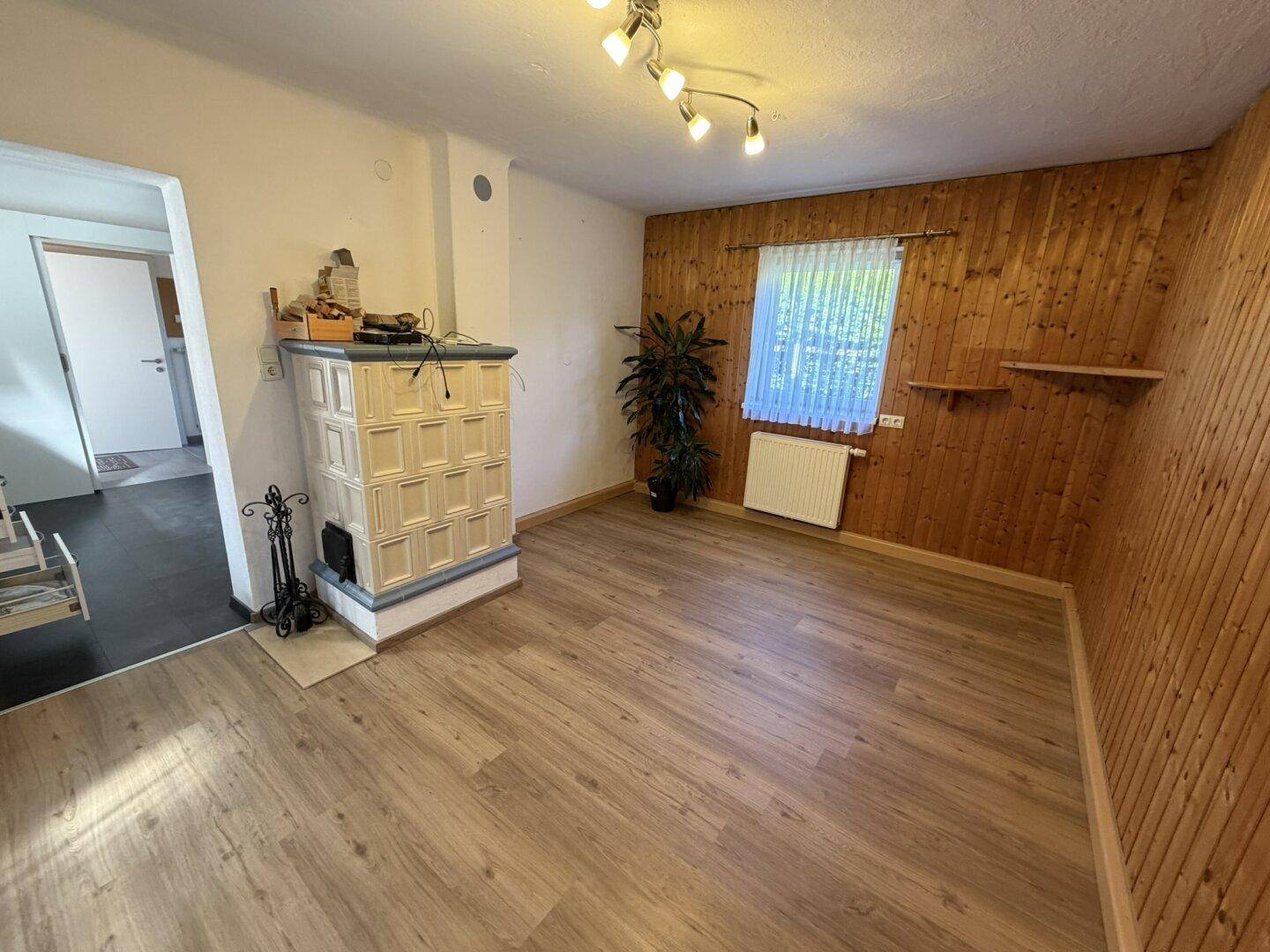 Gemütliches Wohnzimmer mit Holzvertäfelung, Laminatboden und einem traditionellen Kachelofen.