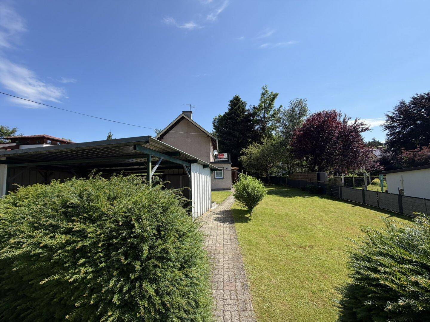 Carport und gepflasterter Weg zum Haus, umgeben von grünen Hecken und Rasen.
