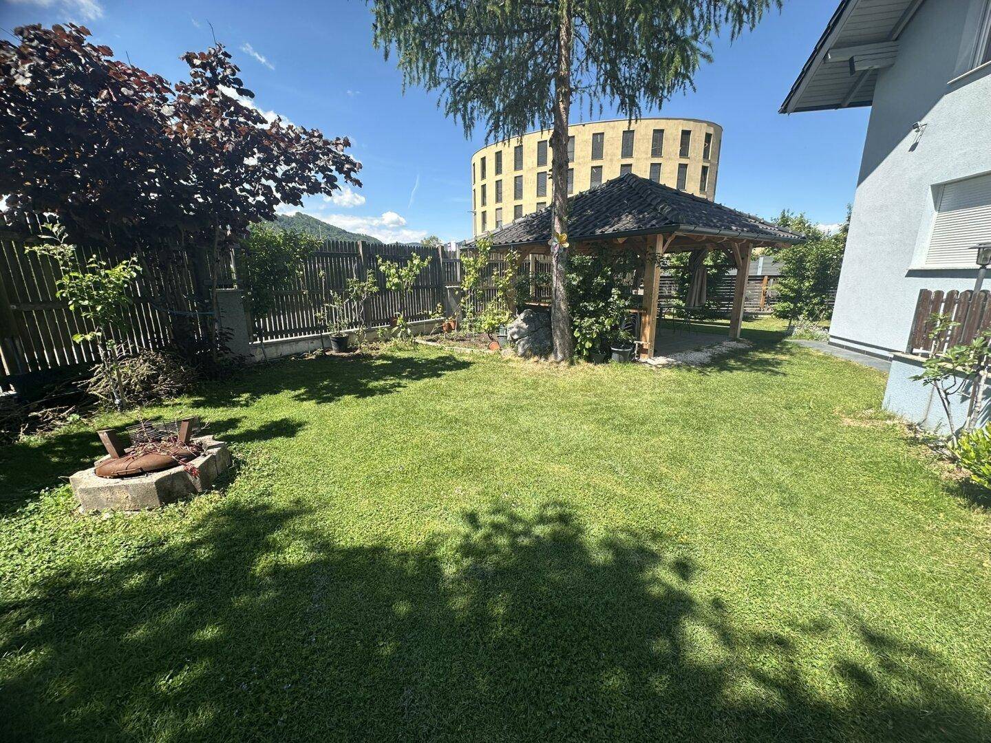 Gepflegter Garten mit Rasenfläche, Gartenlaube und einer Feuerstelle im Freien.