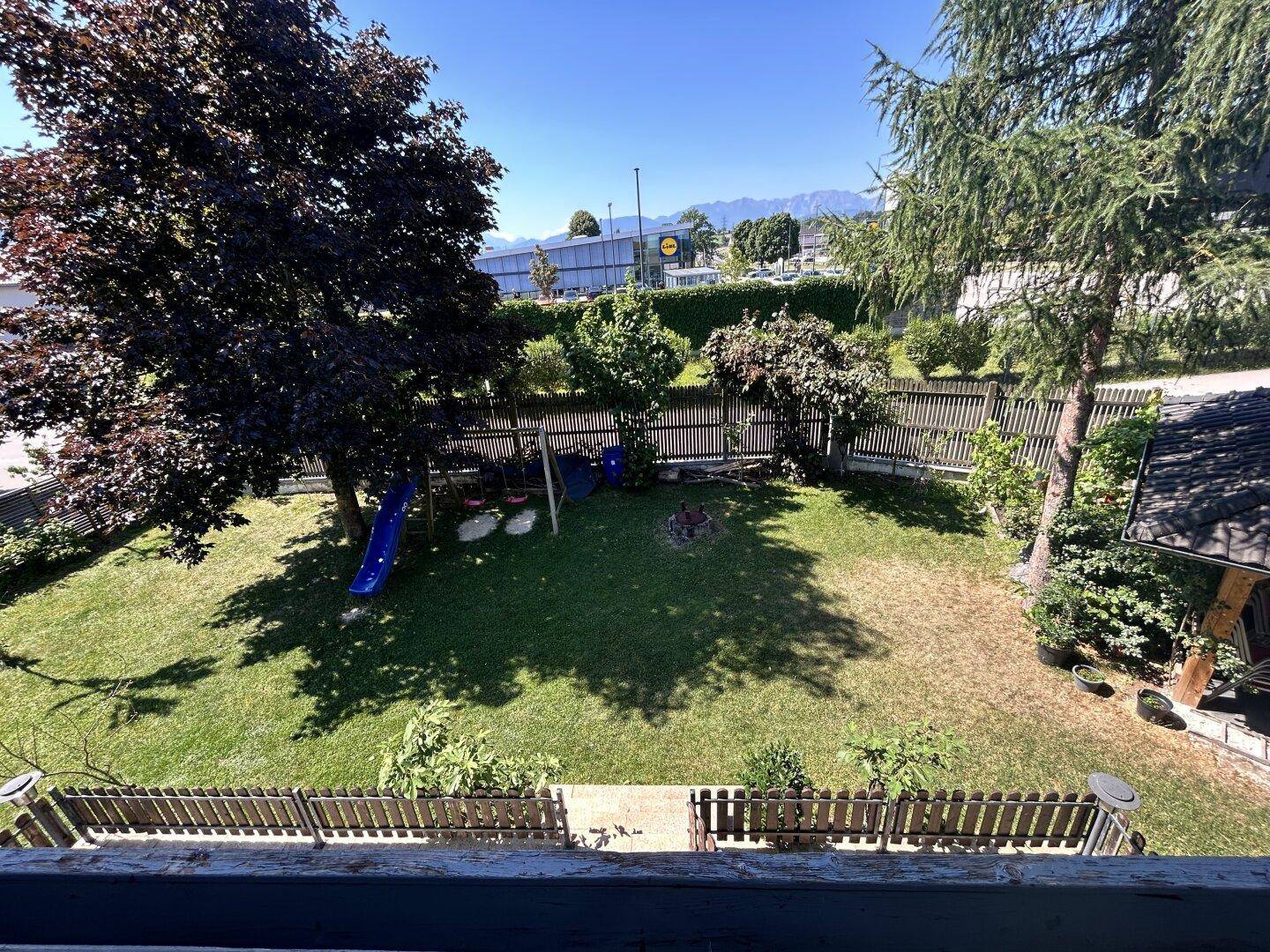 Blick auf den Garten mit Rasenfläche, Spielplatz und hohen Bäumen.