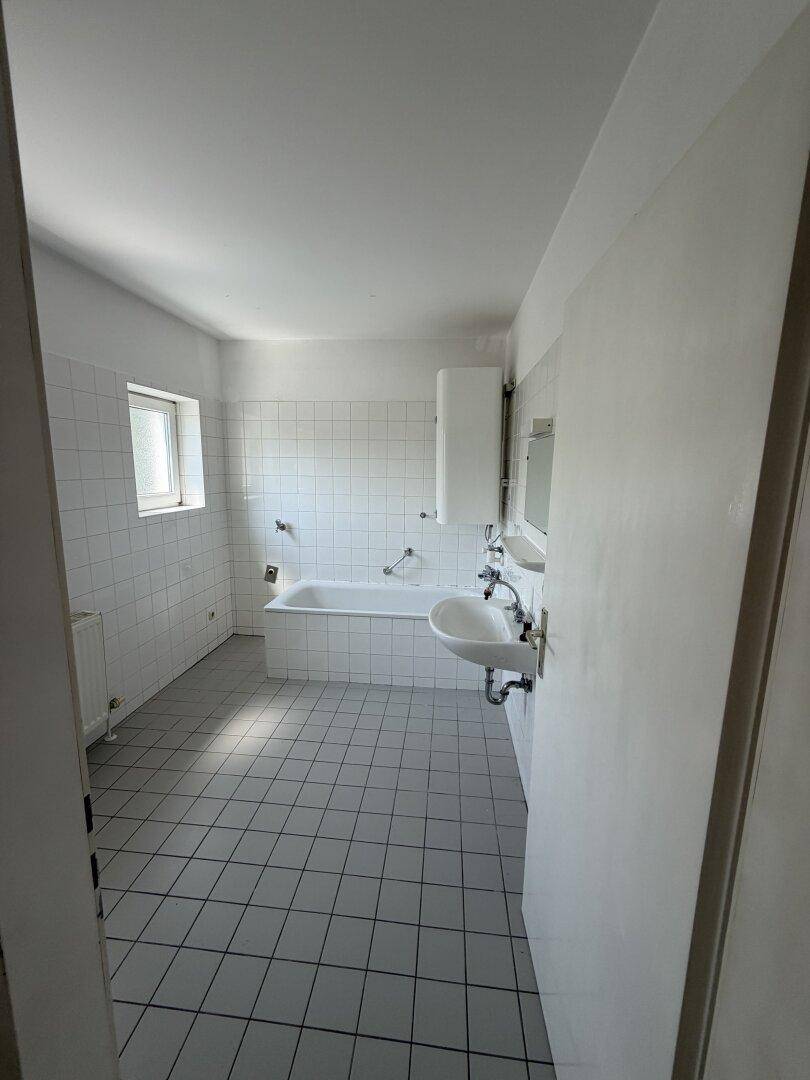 Helles Badezimmer mit weißen Wandfliesen, grauen Bodenfliesen, Badewanne und Fenster.