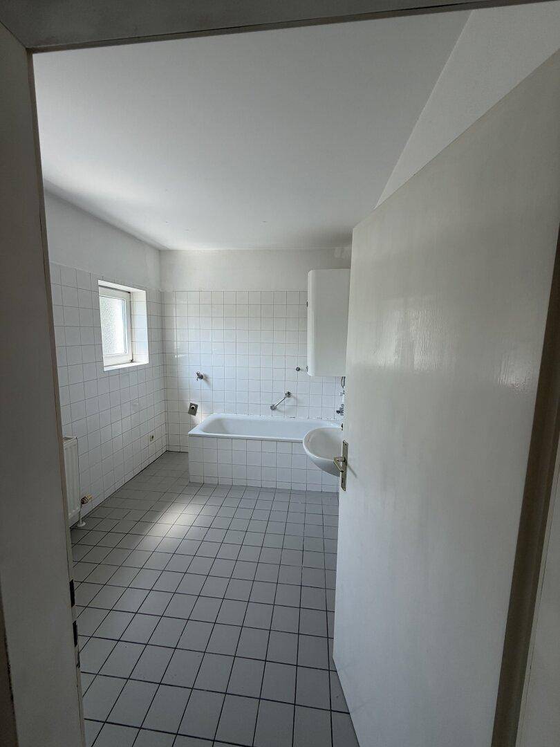 Helles Badezimmer mit weißen Wandfliesen, grauen Bodenfliesen, Badewanne und Fenster.