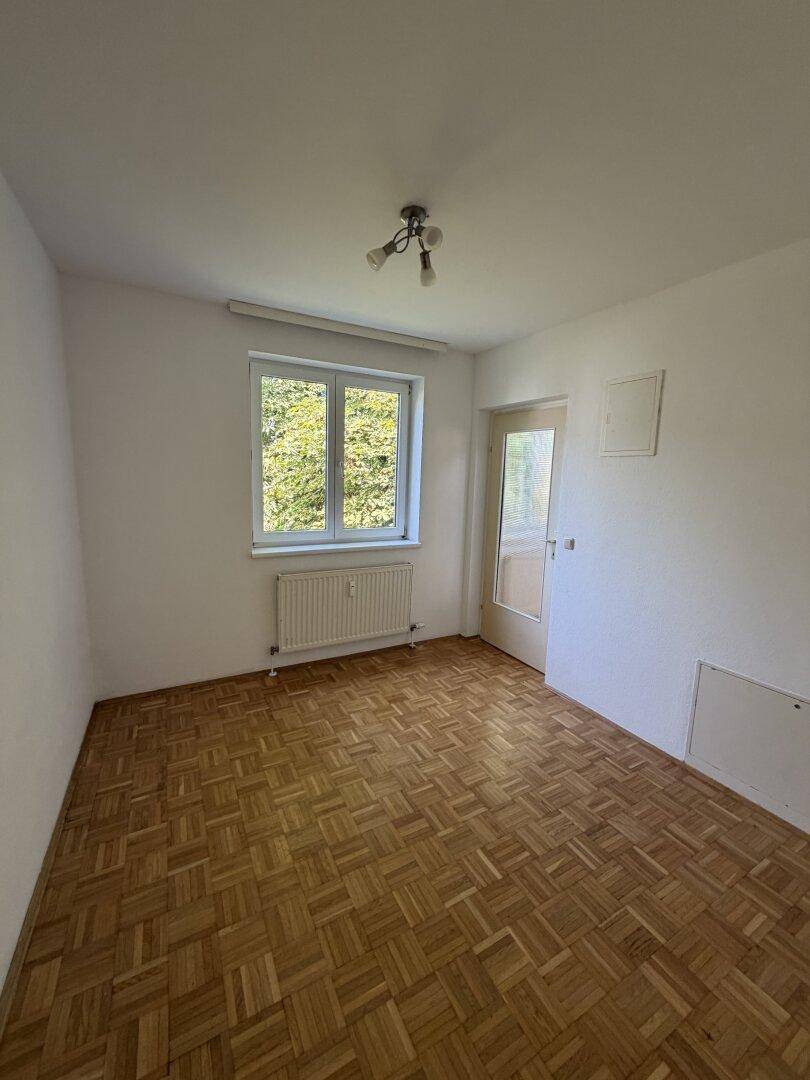 Leeres Schlafzimmer mit Parkettboden, weißen Wänden und einem Fenster mit Blick ins Grüne.