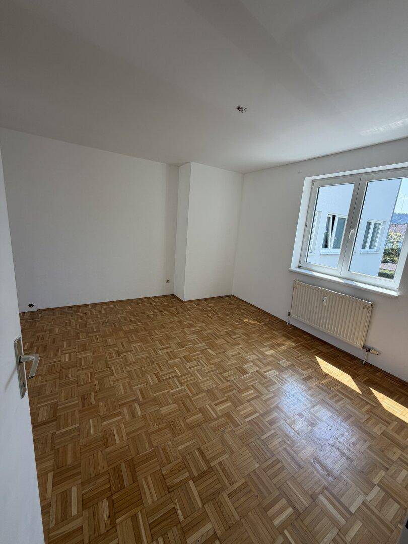 Geräumiges, leeres Zimmer mit Parkettboden und großen Fenstern, die viel Tageslicht hereinlassen.