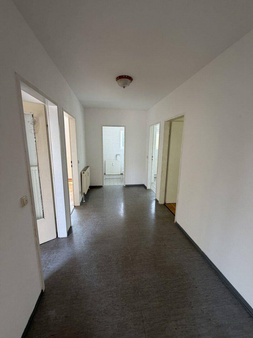 Langer Flur mit dunklem Bodenbelag und weißen Wänden, der zu verschiedenen Räumen führt.