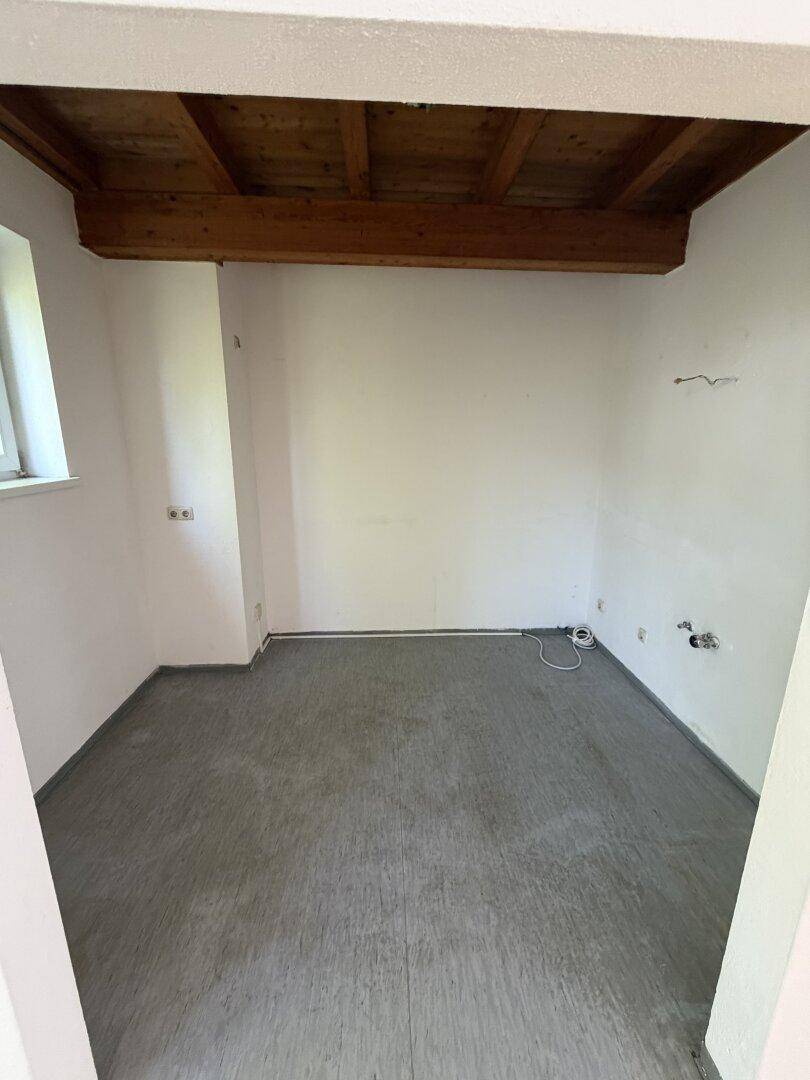 Kleiner, leerer Raum mit dunklem Bodenbelag, weißen Wänden und einem sichtbaren Deckenbalken.