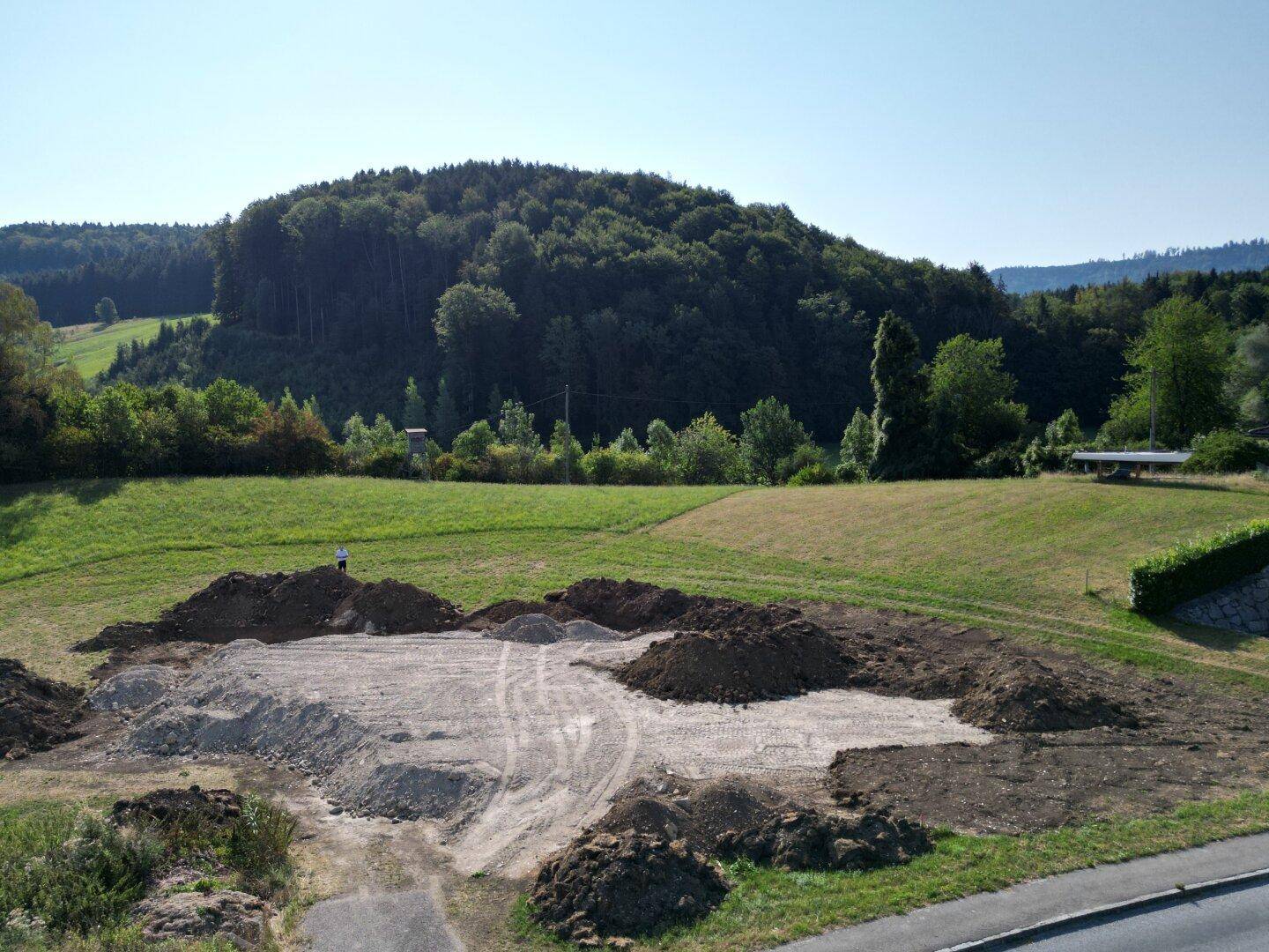 Baugrundstück mit Erdhügeln und vorbereiteter Fläche, umgeben von grünen Wiesen und einem bewaldeten Hügel.