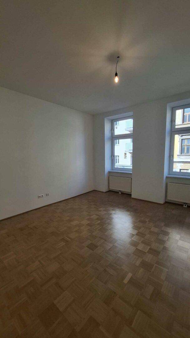 Geräumiges Wohnzimmer mit Parkettboden und zwei großen Fenstern, die viel Licht hereinlassen.
