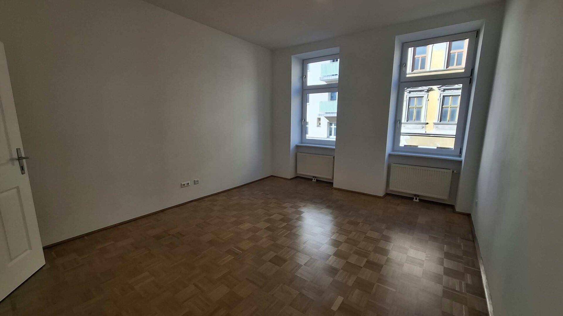 Helles Wohnzimmer mit Parkettboden und zwei Fenstern, die den Raum gut belichten.