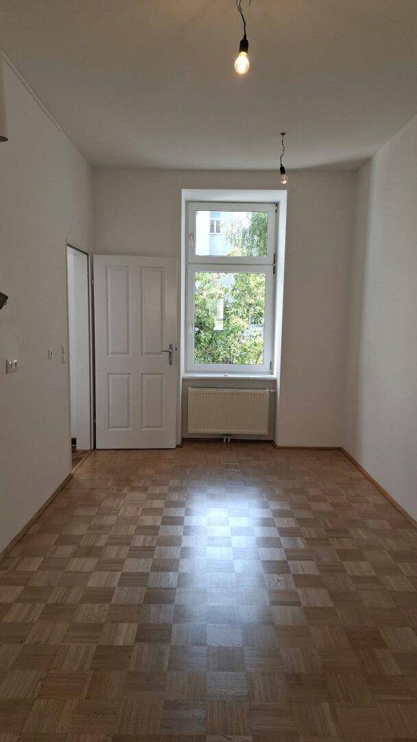 Helles Zimmer mit Parkettboden und einem Fenster, das einen Blick ins Grüne bietet.