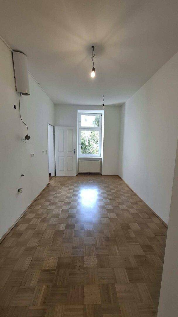 Leeres Zimmer mit Parkettboden und einem Fenster, das den Raum mit Tageslicht versorgt.