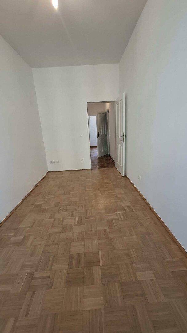 Leeres Zimmer mit Parkettboden und einer Tür, die in einen anderen Bereich der Wohnung führt.