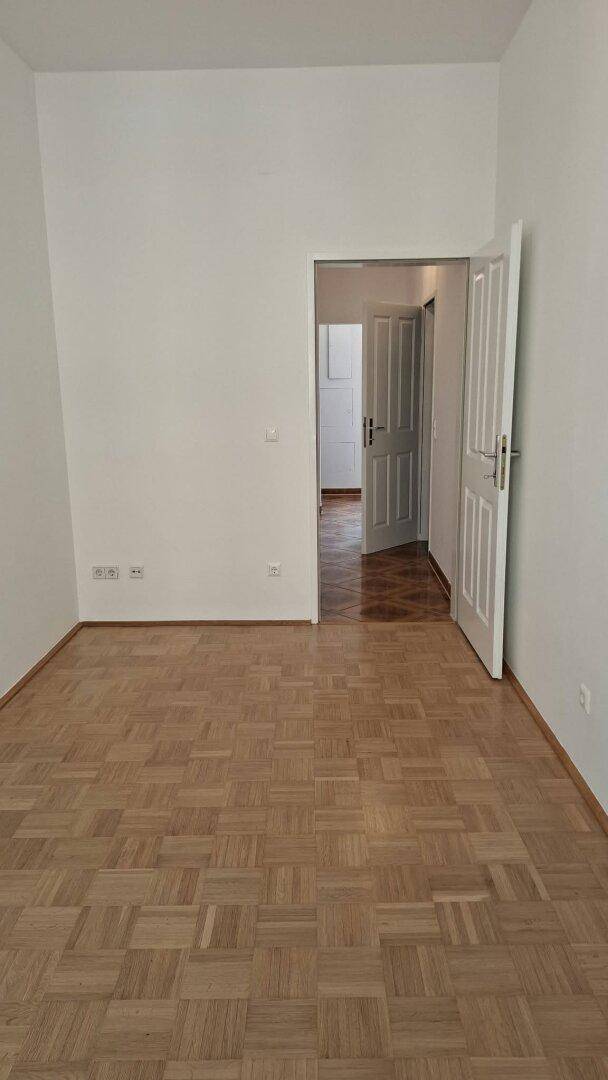 Helles Zimmer mit Parkettboden und einer geöffneten Tür, die den Blick in den Flur freigibt.