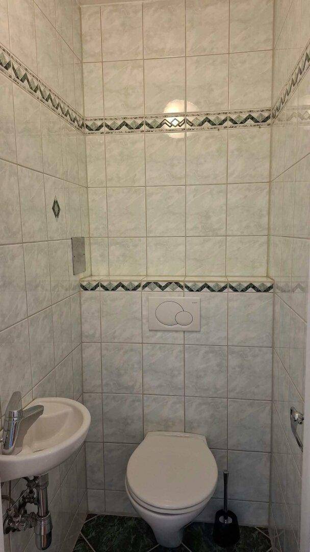 Separate Toilette mit Waschbecken und Wand-WC, gefliest in hellen und dunklen Tönen.