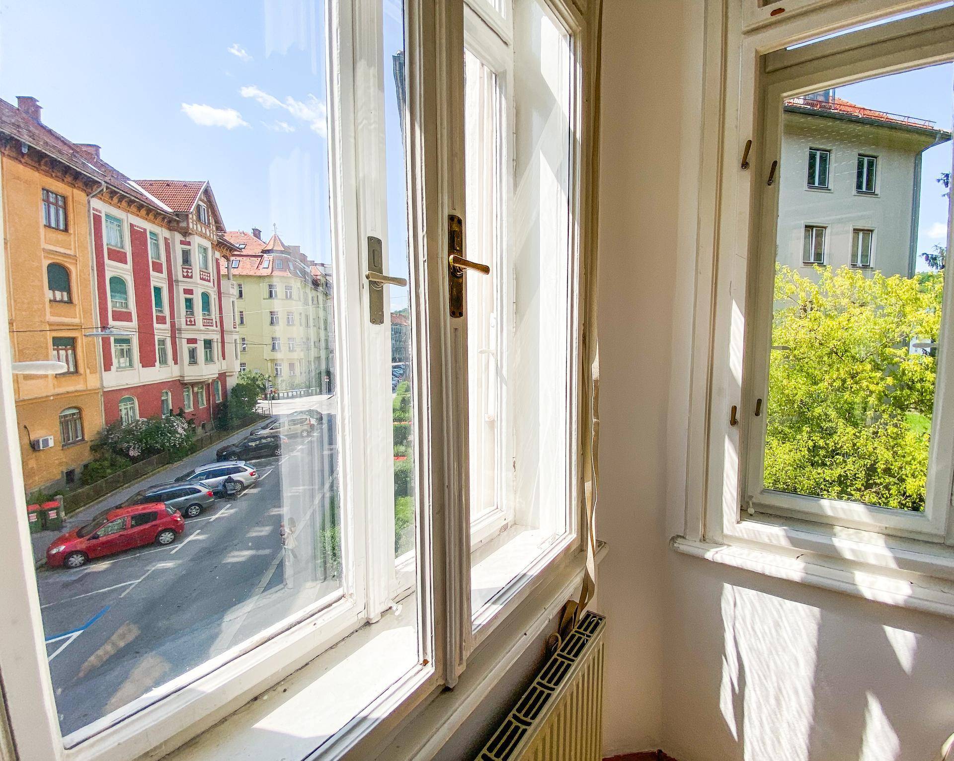 Ausblick aus einem Fenster auf eine belebte Straße mit parkenden Autos und gegenüberliegenden Gebäuden.